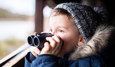 Boy wearing a blue and white knitted wooly hat, and a fur trimmed parka coat. He is stood at the window of a bird hide holding a pair of binoculars to