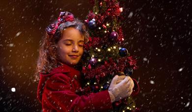 A young girl in a red winter coat hugs a small Christmas tree. She is smiling contentedly with her eyes closed, and has a red tartan bow in her hair.