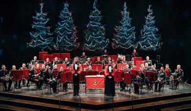 The Band of HM Royal Marines stand on the Festival Theatre Stage dressed in red military jackets, facing out front. Behind them are five blue-lit Chri