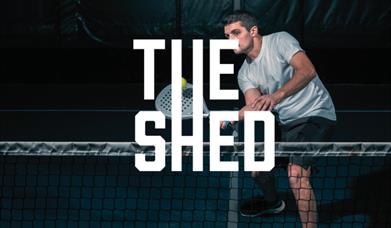 Image of a dark indoor court with a man playing Padel. The logo of The Shed is overlaid onto the image
