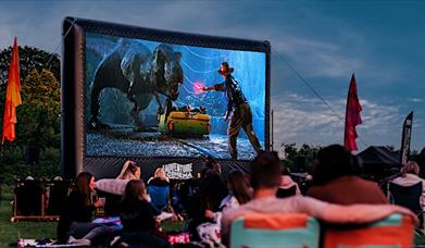 Jurassic Park plays on a large outdoor cinema screen as people picnic in front.