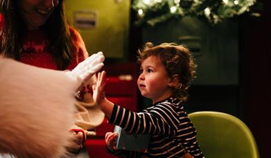 Father Christmas in a red suit with fluffy white trim sits in his grotto with a small child.