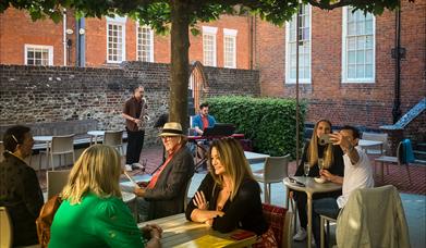 People sit in a sunny evening courtyard enjoying drinks and live music.