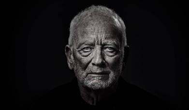 An elderly man (Ian McDiarmid) stares at the camera. His face is wrinkled and looks stern and dramatic, with shadows under his eyes and chin.  He is w