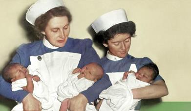 Two midwives in white smocks, white caps and blue blouses stand against a green and white wall. The midwife on the left holds two newborn babies, they