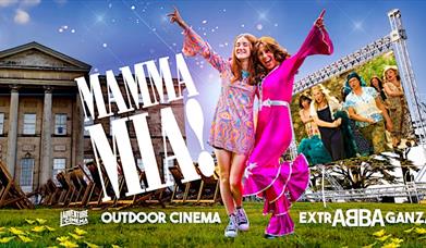 Two women dressed in Abba inspired outfits stand in front of a historic house whilst Mamma Mia plays on a large outdoor cinema screen.