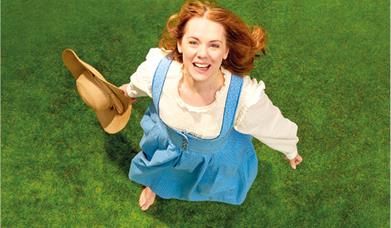 A woman in a blue dress and white blouse is seen from above. She looks up at the camera smiling. Her arms are spread slightly, and she holds a straw h