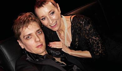 A young man and an older woman are lit up as though by a paparazzi camera. He is dressed in a black silk shirt and looks at the camera, the woman lean